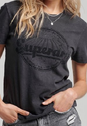 Frau trägt ein dunkelgraues Superdry T-Shirt mit funkelndem Logo, kombiniert mit zerrissenen hellgrauen Jeans, Hände in den Taschen.
