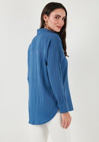 Camisa azul con textura, de botones, con cuello y mangas largas, que presenta un dobladillo curvado y sutiles rayas verticales, combinada con pantalones blancos.