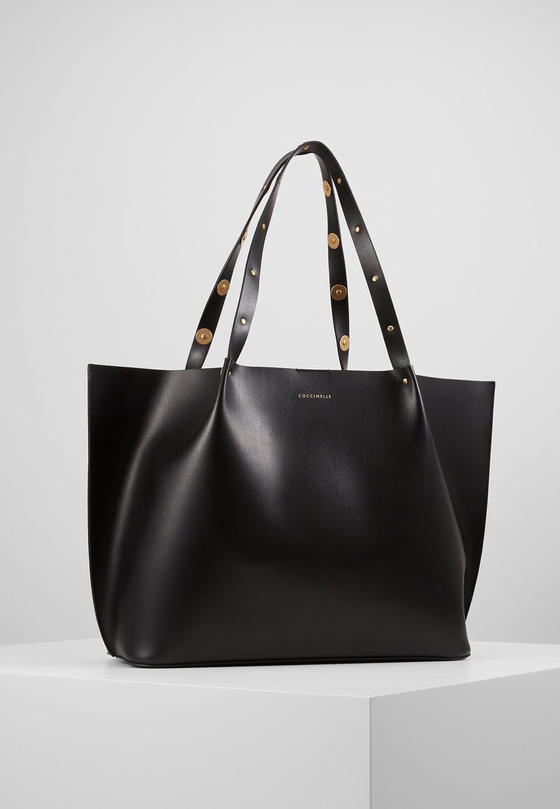 Sac cabas en cuir noir avec des clous dorés sur les doubles anses, corps légèrement froissé et petit logo de la marque à l’avant, présenté sur une surface blanche.