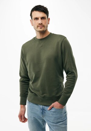 Mężczyzna w oliwkowozielonej bluzie z długim rękawem i jasnoniebieskich dżinsach, stojący z jedną ręką w kieszeni na białym tle.