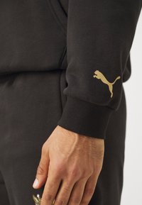 Hanorac negru cu un logo auriu Puma pe manșon. Țesătura pare moale, cu un mansetă cu nervuri, evidențiind o potrivire strânsă.