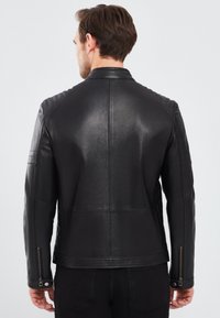 Zwarte leren jas met een aangesloten ontwerp, gestikte schouderaccenten, ritsmanchetten en een gladde textuur. Achteraanzicht toont stikseldetails.