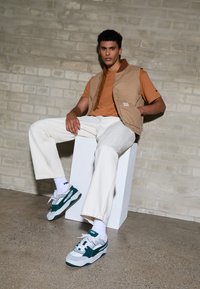 Gilet color tabacco sopra una maglietta arancione, pantaloni bianchi a gamba larga e sneakers teal/grigie. La superficie è liscia con una texture opaca. Sullo sfondo c'è un muro di mattoni.