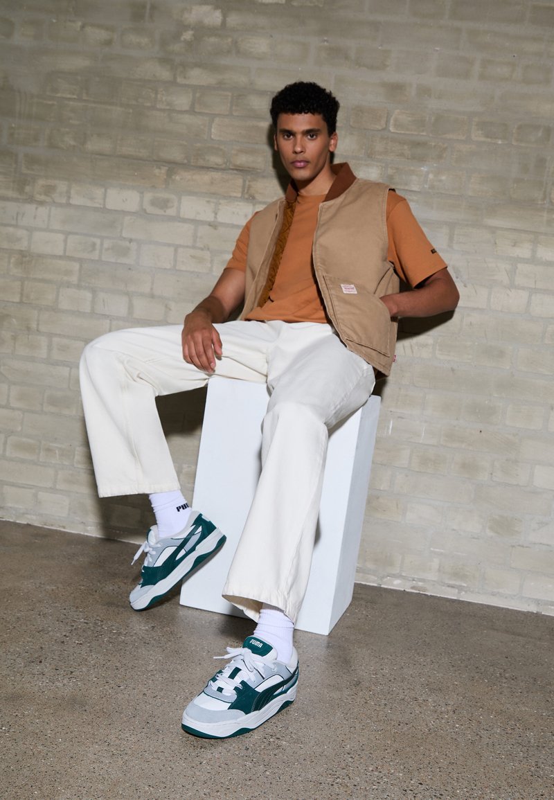 Gilet color tabacco sopra una maglietta arancione, pantaloni bianchi a gamba larga e sneakers teal/grigie. La superficie è liscia con una texture opaca. Sullo sfondo c'è un muro di mattoni.