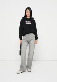 Sweatshirt preto com capuz com o texto "TOMMY JEANS" em rosa, combinado com jeans largos cinzentos e sapatos beges, segurando uma bolsa preta.