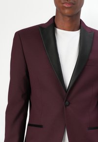 Burgunderfarbener Blazer mit schwarzen Satinrevers, ein Knopfverschluss und zwei Vorderaschen. Glatte Textur mit strukturierter Silhouette.