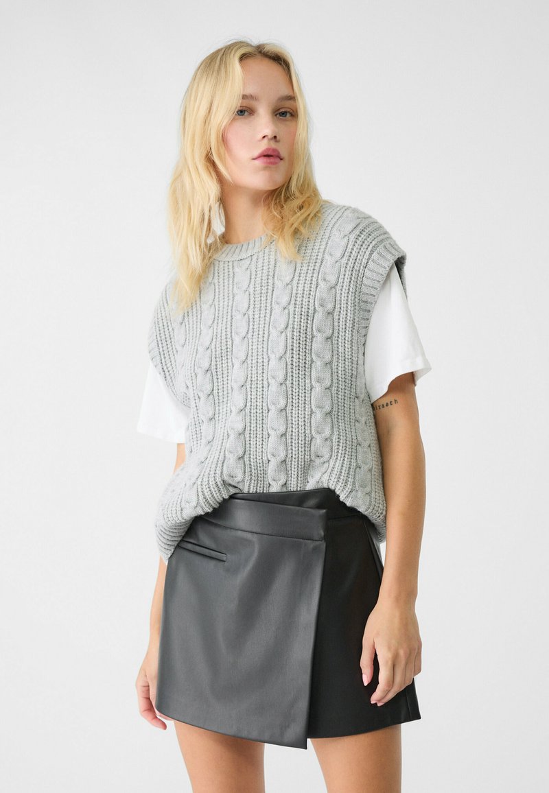 Donna bionda che indossa un maglione senza maniche grigio a trecce sopra una camicia bianca a maniche corte e una minigonna in pelle nera asimmetrica.