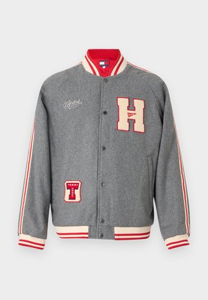 Veste universitaire grise avec des accents rouges et crème, ornée de broderies de patches 'H' et 'TOMMY JEANS'. Col et poignets côtelés. Fermeture à boutons-pression.