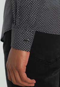 Czarna i biała koszula w wzór z rękawem na mankiet, w diamentowe kształty, z zapięciem na guzik i gładką fakturą materiału, noszona na ciemnych spodniach.