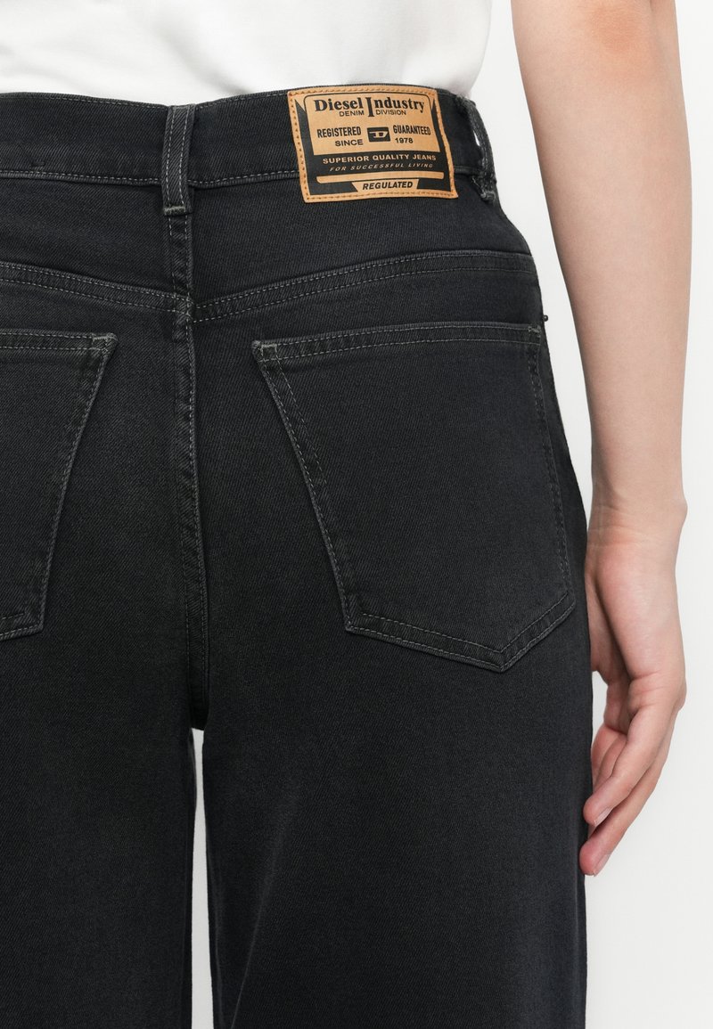 Jeans en denim noir avec une texture lisse, présentant une étiquette en cuir marron au niveau de la taille et deux poches arrière avec des coutures soignées.