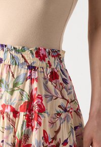 Gonna maxi floreale con base beige, caratterizzata da vivaci motivi rossi, blu e verdi. È visibile una vita elastica con bordi arricciati.