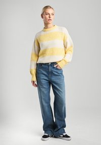 Keltainen ja valkoinen raidallinen neulepaita pitkillä hihoilla, yhdistettynä korkeavyötäröisiin vaaleansinisiin leveälahkeisiin farkkuihin ja mustiin lenkkareihin.