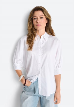 Frau mit welligem Haar, die ein lockeres weißes Hemd mit Puffärmeln und hellblaue Jeans trägt, steht mit einer Hand in der Tasche.