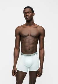 Αθλητικός άνδρας με σκούρο δέρμα, που φοράει λευκά boxer briefs Calvin Klein, στέκεται μπροστά σε απλό φωτεινό φόντο, κοιτάζοντας μπροστά με ουδέτερη έκφραση.