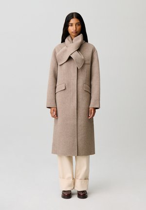 Donna che indossa un lungo cappotto di lana beige con sciarpa integrata, pantaloni color crema a gamba larga e scarpe marrone scuro, in piedi davanti a uno sfondo semplice.