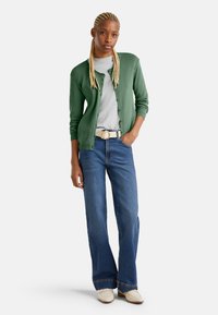 Zelený svetr přes šedou košili, kombinovaný s rozšířenými modrými džínami a světlými botami. Výrazná kulatá spona opasku a strukturovaný svetr.