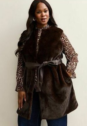 Braune Fleece-Weste mit Bindegürtel, über einem gemusterten braunen Oberteil getragen, das lange Ärmel und einen lockeren Schnitt hat.