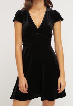 Femme portant une robe courte en velours noir à manches courtes, avec un col en V et une taille cintrée, debout devant un fond uni.