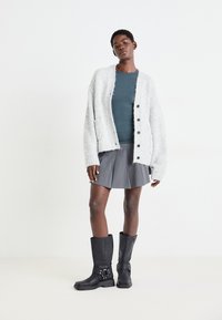 Cardigan lavorato a maglia di un grigio chiaro con tasche frontali e bottoni, abbinato a un top teal, una gonna plissettata grigia e stivali neri fino al ginocchio con fibbie.