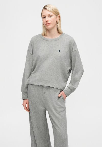 Ξανθιά γυναίκα με ανοιχτόχρωμο γκρι μακρυμάνικο κορμάκι και ταιριαστό άνετο παντελόνι, στέκεται με το ένα χέρι στην τσέπη, μπροστά σε λευκό φόντο.