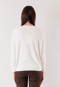 Maglione bianco a maniche lunghe, collo rotondo e un'etichetta visibile sul retro. Texture liscia e vestibilità comoda; abbinato a pantaloni marroni.