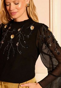 Črna bluzica z teksturirano tkanino, ki ima cvetlične aplikacije iz sequins in prosojne rokave. Zlati okrogli uhani dopolnjujejo dizajn.