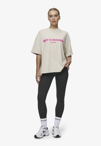 Beige Übergröße T-Shirt mit pinkem Text "SLEEP IS OVERRATED", kombiniert mit schwarzen Leggings und weißen Turnschuhen. Lässige und entspannte Passform.