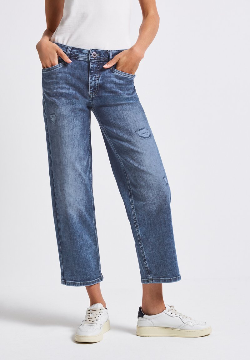Jeans en denim bleu clair avec une coupe décontractée, dotés de poches avant, de détails déchirés et d'ourlets retroussés, associés à des baskets blanches.