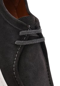 Zapato de ante negro con un acabado texturizado, que presenta cordones grises y un contraste en las costuras. El diseño incluye una silueta de perfil bajo y material suave.