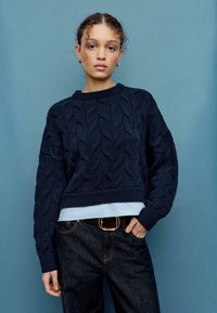 Maglione blu navy a trecce con design corto e bordi a coste, indossato sopra una camicia azzurro chiaro, abbinato a jeans di denim scuro.