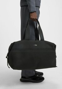 Sac de sport noir en tissu texturé avec poignées doubles, muni d'une fermeture éclair et du logo "BOSS" à l'avant. Design compact et structuré.