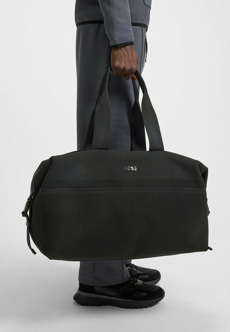 Sac de sport noir en tissu texturé avec poignées doubles, muni d'une fermeture éclair et du logo "BOSS" à l'avant. Design compact et structuré.