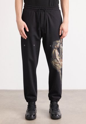 Black sweatpants with a drawstring waist, tapered cuffs, and a graphic design on the left thigh featuring a colorful figure.