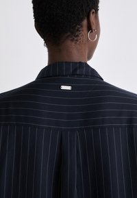 Navy striped shirt with a back pleat, featuring a smooth texture and a silver hardware accent at the collar.