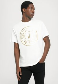 Λευκό βαμβακερό t-shirt με χρυσό κυκλικό λογότυπο που έχει το κείμενο "VERSACE" και "COUTURE". Έχει κοντά μανίκια και άνετη εφαρμογή.