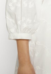 Bílá vyšívaná blůza s balónovými rukávy, květinovými vzory a ohrnutým manžetem. Tkanina má měkký, strukturovaný vzhled.