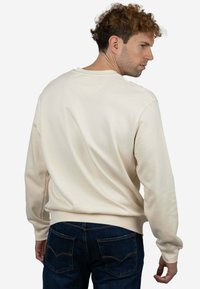 Sudadera color crema con mangas largas y cuello redondeado, con una textura suave. Combinada con vaqueros de mezclilla azul oscuro.