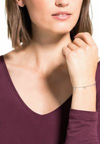 Pulsera de plata con pequeños acentos circulares, llevada en una muñeca de tono de piel claro. Fondo con una blusa burdeos de escote en V profundo.