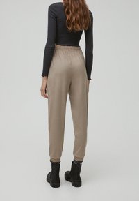 En kvinna med långt, vågigt hår bär en svart croptop och beige joggers, stående mot en ljusgrå bakgrund, med fokus på sin outfit.