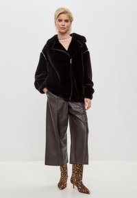 Zwarte nepbont jas met rits aan de voorkant, brede mouwen en grote kraag, gecombineerd met hoogwaisted grijze leren broek en enkelboots met luipaardprint.