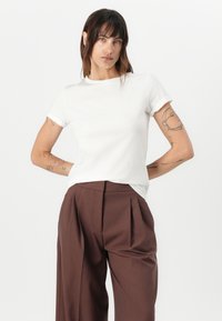 White cotton t-shirt with short sleeves, round neckline, and a relaxed fit, paired with brown pleated trousers. Minimalist design, no visible patterns.