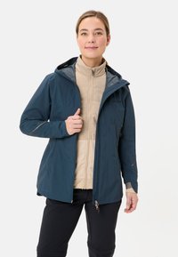 Marineblauwe waterdichte jas met een gevoerde capuchon, rits aan de voorkant en elastische manchetten, gedragen over een lichtgewicht beige binnenlaag, tegen een effen achtergrond.