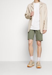 Chaqueta ligera beige sobre una camiseta blanca, combinada con pantalones cortos verde oliva y zapatillas deportivas beige con acentos naranjas; calcetines blancos visibles.