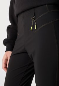 Black athletic pants featuring zippered pockets, green accents on drawstrings, and contrast stitching for added detail.