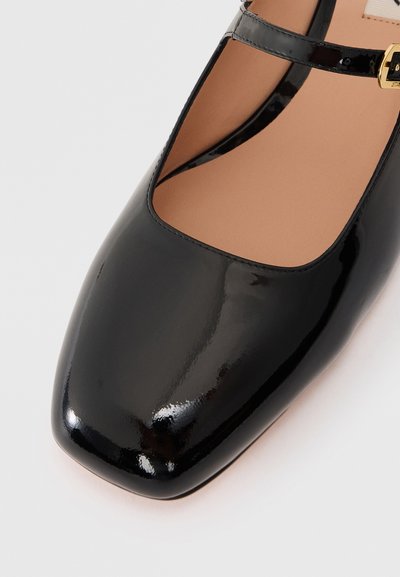 Chaussure en cuir verni noir avec un bout carré arrondi et une fine bride de cheville. La semelle est marron clair, offrant une finition élégante.