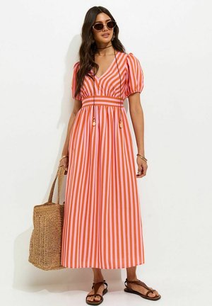 Femme portant une robe longue à manches bouffantes rayée orange et rose, des lunettes de soleil, des sandales noires, et tenant un grand sac fourre-tout tissé.