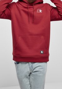 Sweat à capuche bordeaux en tissu doux, avec une poche kangourou à l'avant, un logo étoile blanc sur la poitrine, et des poignets et ourlet côtelés.