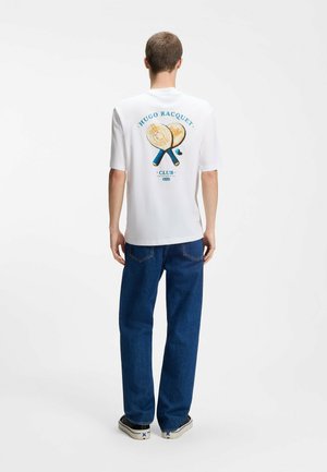 Man draagt een wit t-shirt met gekruiste houten rackets en tekst "Hugo Racquet Club" op de rug, gecombineerd met een blauwe spijkerbroek en sneakers.