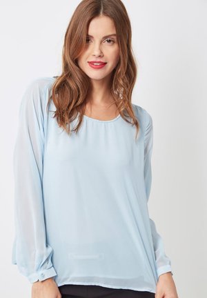 Mujer con cabello largo castaño, vistiendo una blusa de manga larga azul claro y pantalones negros, sonriendo frente a un fondo blanco liso.