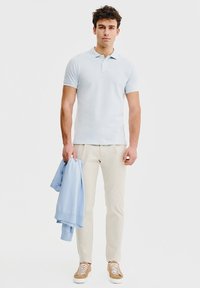 Jasnoniebieska koszulka polo, beżowe spodnie, trzymający jasnoniebieski sweter. Sneakersy z białymi podeszwami, krótkie rękawy, gładka tekstura materiału.